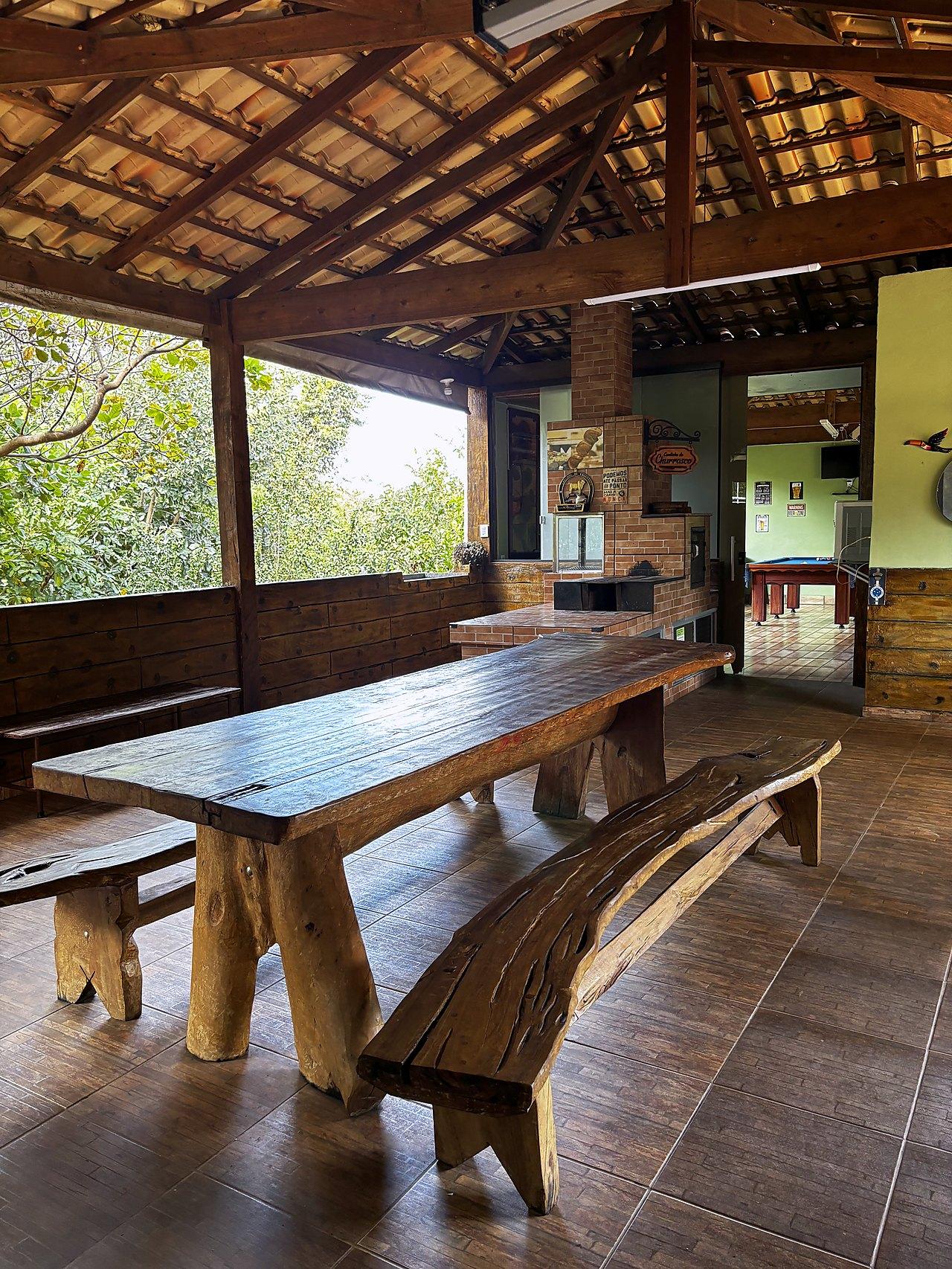 Rancho coberto com mesa rústica de madeira e churrasqueira ao fundo